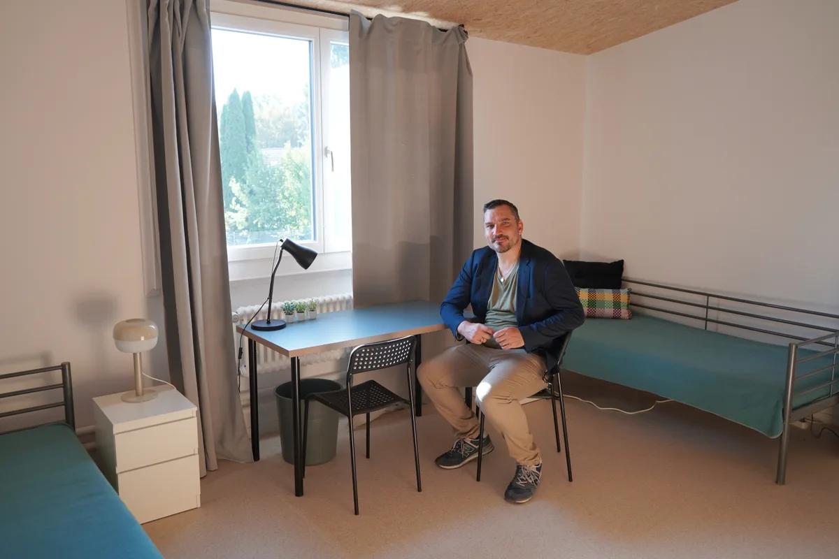 Ein Mann sitzt in einem Zweibettzimmer an einem Pult und schaut in die Kamera. 